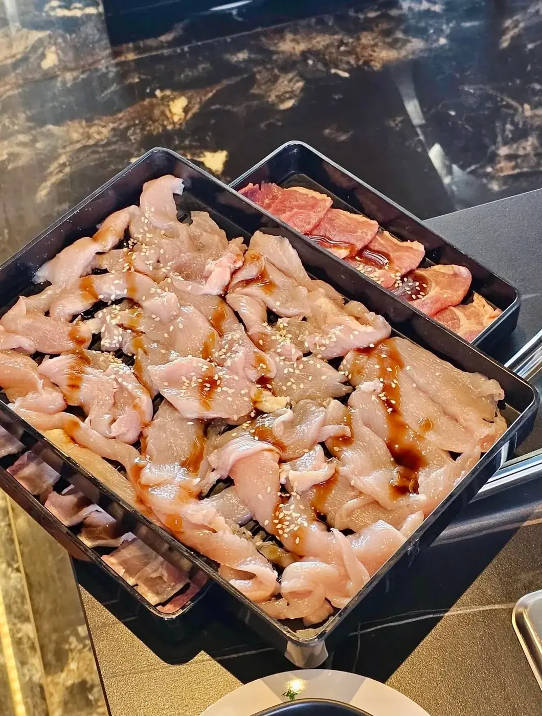 Trays of thinly sliced chicken and pork, glazed and scattered with sesame, ready to cook