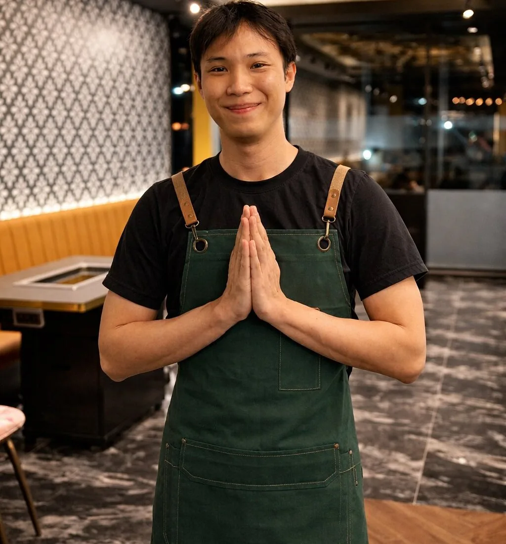 A smiling member of the Mama Suki team in a green apron, greeting guests with his palms together