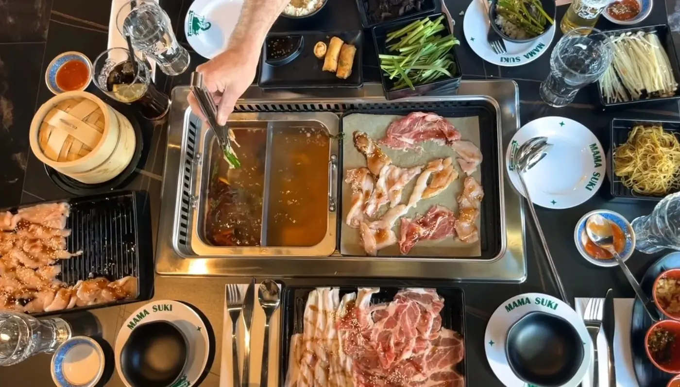 A Mama Suki table from above: a split pot of two broths bubbling beside the grill, surrounded by sliced meats, noodles, greens and dipping sauces
